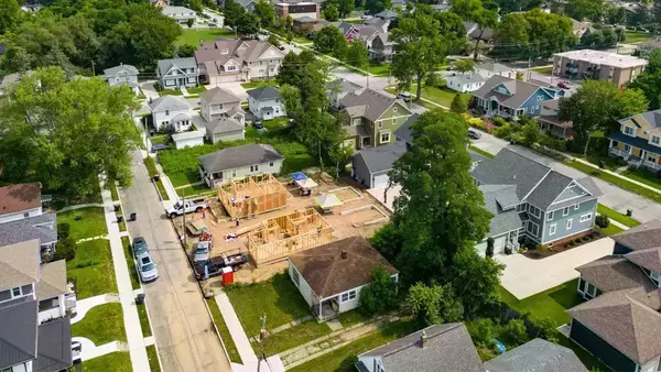 Aerial view of a residential neighborhood with a new home construction site. The wooden frame of the house is visible, surrounded by construction materials and equipment.  Completed houses of various sizes and styles line the paved streets, interspersed with green lawns and trees.