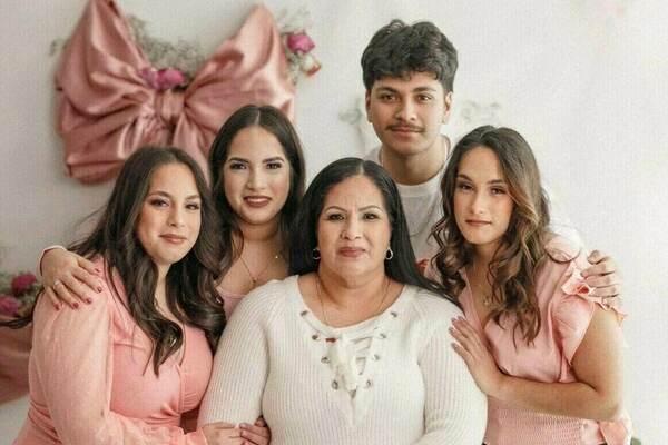 A family of five poses for a photo. A woman sits in the center wearing a white sweater and jeans, flanked by two young women in pink tops and dark pants.  A young man stands behind the seated woman, and another young woman stands behind the woman on the right. A large pink bow decorates the wall behind them.