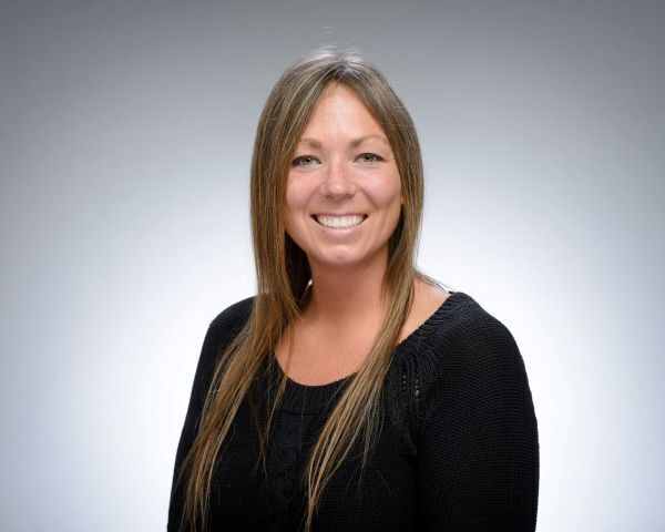 Smiling woman with long, straight brown hair and green eyes, wearing a black textured sweater against a gray background.